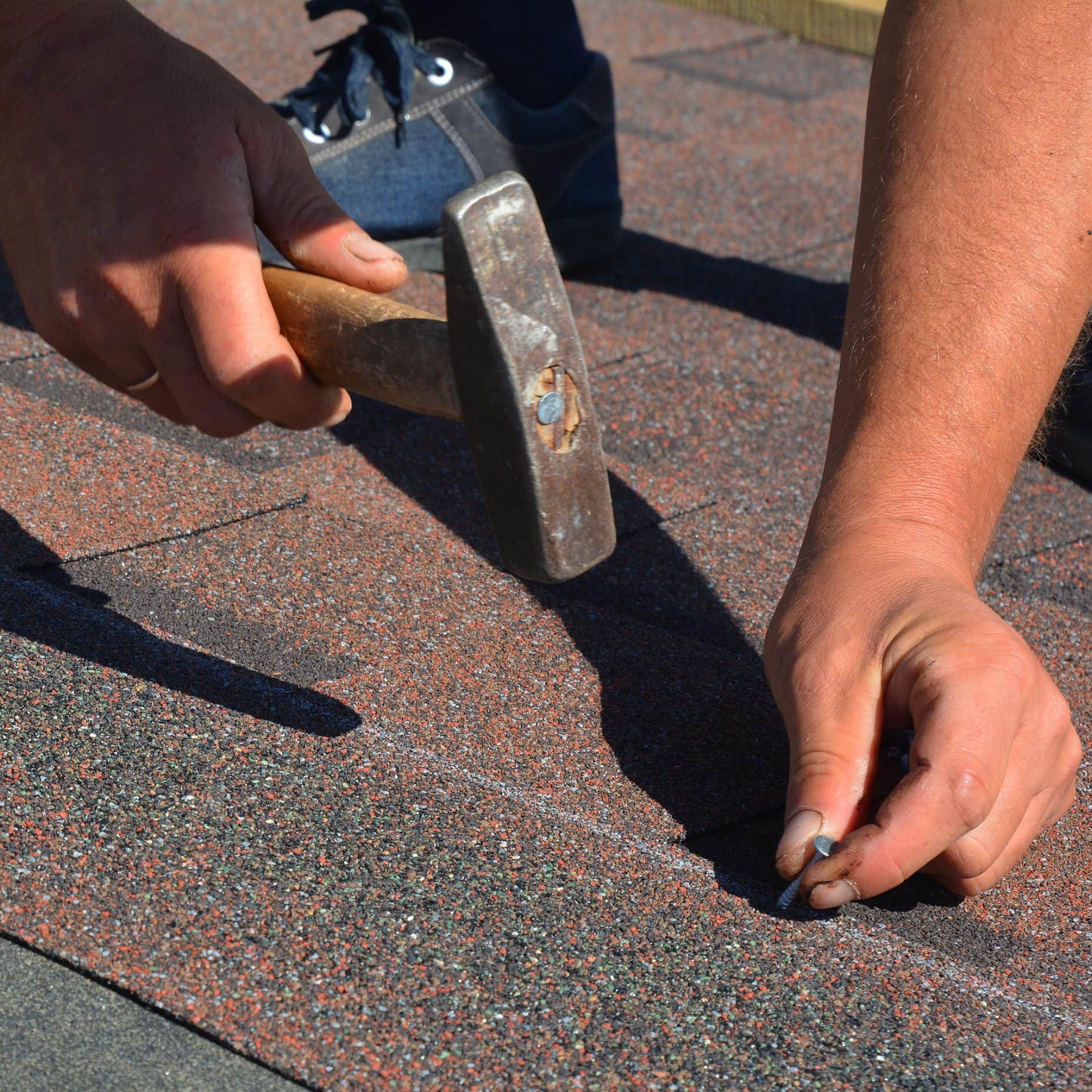Worker installing asphalt shingles on residential roof with hammer and nails.